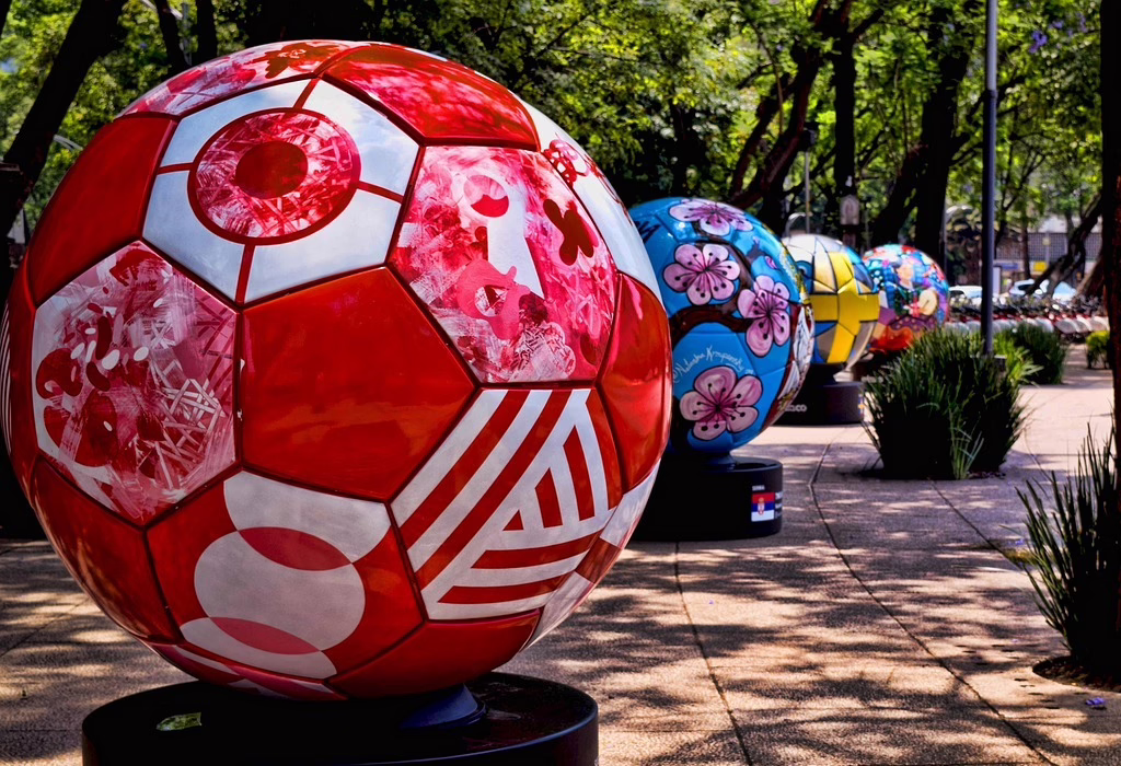 A row of large, colorful painted football / soccer sculptures on a sunny pedestrian path with green trees in Mexico City.